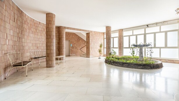 Eine geräumige Lobby mit gewölbter Ziegelwand, einer zentralen Säule und einer Sitzecke mit Metallstühlen.
