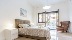 A bedroom with a bed, nightstand, chair, and a window with a view of buildings. - GuestHost - Vomero Deluxe Paris with terrace (Napoli)