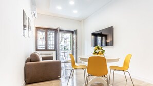 A modern living room with a sofa, a dining table with chairs, a flat-screen TV mounted on the wall, and a large window. - GuestHost - Vomero Deluxe Paris with terrace (Napoli)