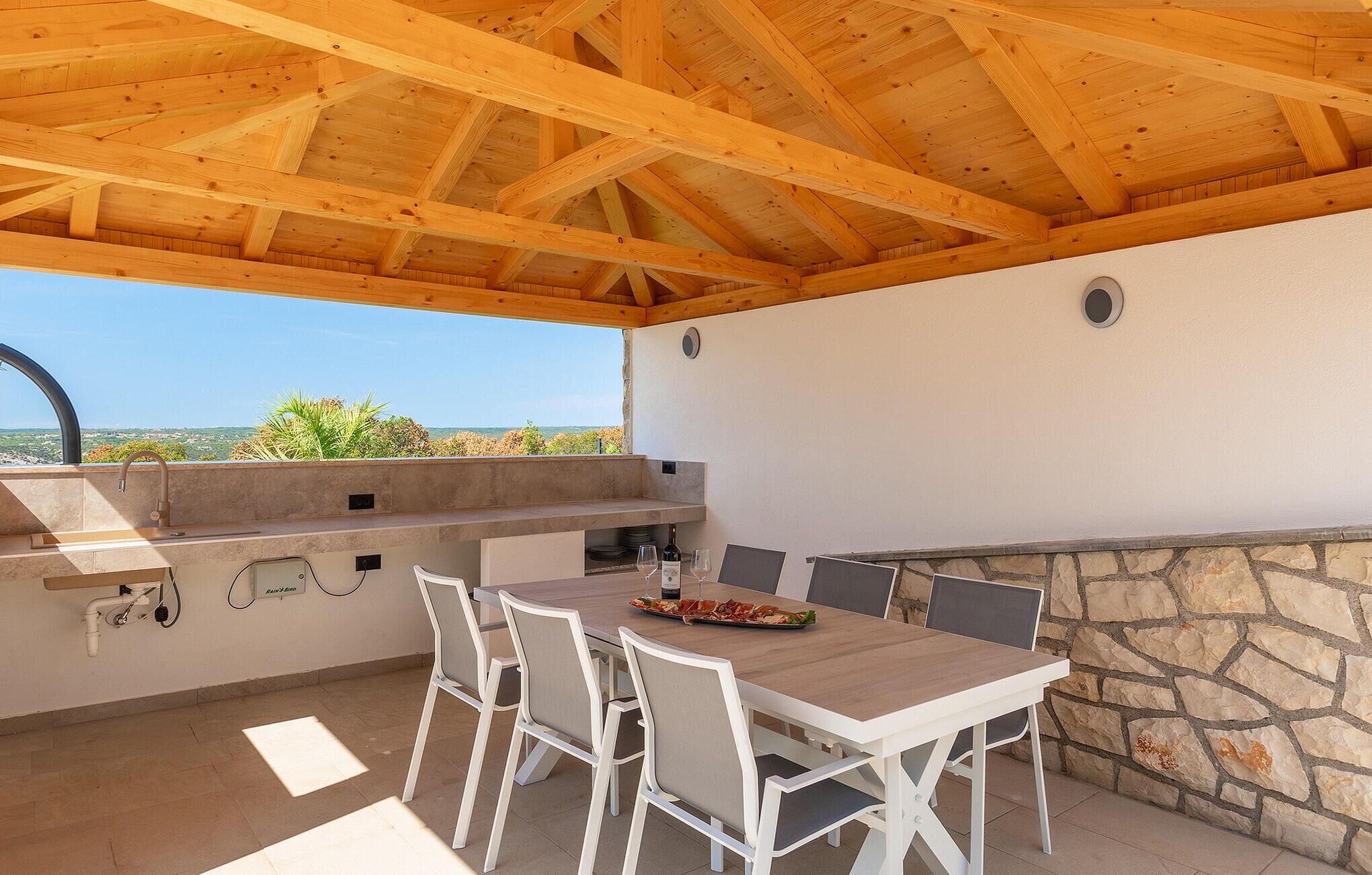 An outdoor patio with a stone wall, a wooden ceiling, and a table with chairs.