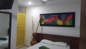 Ein Hotelzimmer mit einem Bett, einem Deckenventilator, einem farbenfrohen abstrakten Gemälde, einer Pflanze in einer Vase und einer gelben Tür.