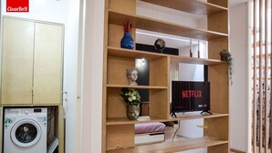 Habitaci ón con lavadora, televisión con Netflix, estanterías de madera y una planta pequeña.