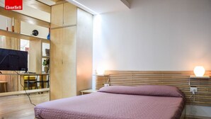 Un dormitorio con cama, cabecero de madera, lámpara de noche y televisor sobre una estantería de madera.