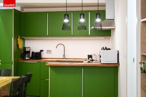 Une cuisine avec des meubles de couleur verte, un plan de travail en bois, un évier, une machine à café, un four à micro-ondes et des luminaires suspendus.