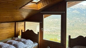 Una habitación con paredes y techo de madera, dos camas con ropa blanca, un ventanal con vistas a un valle y un tragaluz.