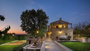 Une maison à deux étages avec une façade en pierre, un court de tennis et un espace extérieur bien éclairé.