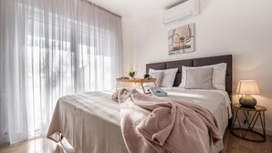 A bedroom with a bed, a bedside table with a lamp, a window with sheer curtains, and an air conditioning unit on the wall.