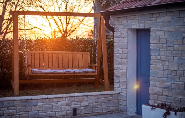 A wooden swing with a cushion, set against a stone wall and a blue door, with a sunset in the background.
