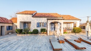A two-story house with a tiled roof, a covered patio with lounge chairs, and a small garden area.
