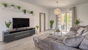 Ein modernes Wohnzimmer mit grauem Sofa, einem Flachbildfernseher, einem Konsolentisch mit Zimmerpflanzen und einem großen Fenster mit Vorhängen.