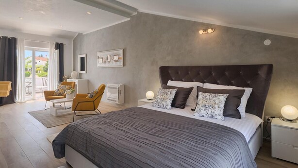 Ein modernes Schlafzimmer mit einem großen Bett, einer Couch und einem Sessel. Ein Fenster mit Vorhängen, ein Heizkörper und eine Wandleuchte sind ebenfalls vorhanden.