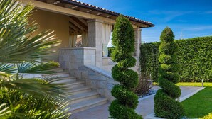 Una casa con una escalera de piedra que conduce a un porche cubierto, rodeada de setos cuidados y un césped.
