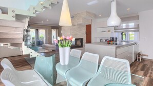 Una cocina moderna con una isla central, un comedor con sillas blancas y una escalera con barandillas de vidrio.