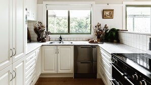 A kitchen with white cabinets, a stainless steel dishwasher, a black stove, and a window with a view of greenery outside. - Cosy Retreat with Mountain Views (Warburton)