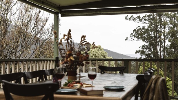 A wooden table with chairs on a balcony, a vase with dried flowers, and a glass of red wine. - Cosy Retreat with Mountain Views (Warburton)