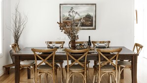 A dining area with a wooden table set for four, surrounded by wicker chairs. A vase with dried branches and a painting of a rural scene are on the wall above the table. - Cosy Retreat with Mountain Views (Warburton)
