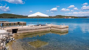 Klares Wasserufer mit einer Betonsteg, einem weißen Sonnenschirm und einem ruhigen blauen Meer.