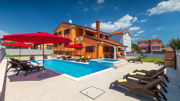 Zona de piscina con sillas de descanso y sombrillas, un edificio de dos pisos con tejado de tejas rojas y un cielo azul despejado.