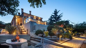 Une maison moderne avec une façade en pierre, un escalier menant à un balcon et un espace extérieur bien éclairé, aménagé avec une table et un banc.