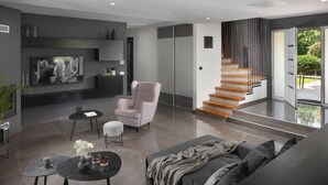 Un salon moderne avec un canapé gris, un fauteuil rose et une table basse noire. Un escalier mène à un balcon d’où l’on peut admirer la verdure.