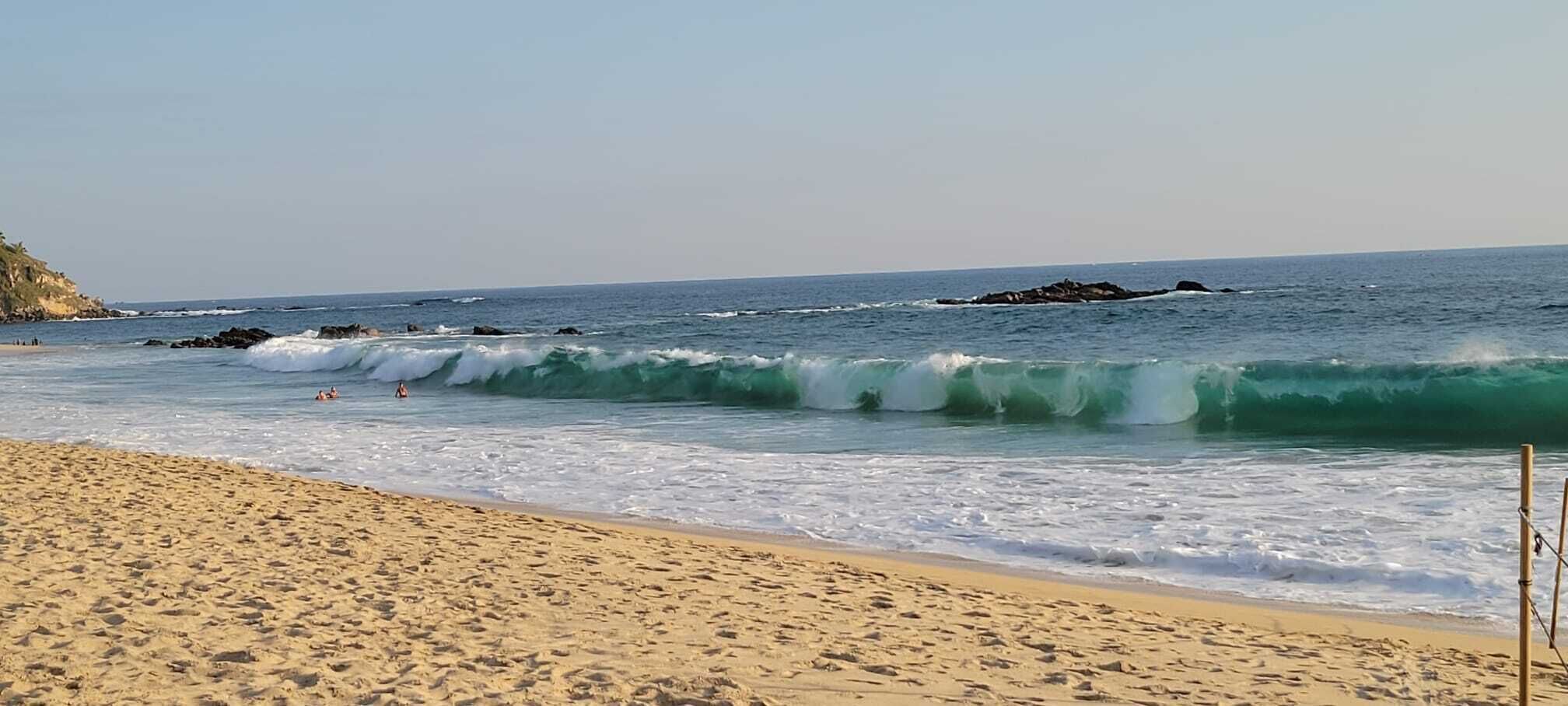 A sandy beach with waves, a clear sky, and rocky outcrops in the distance.