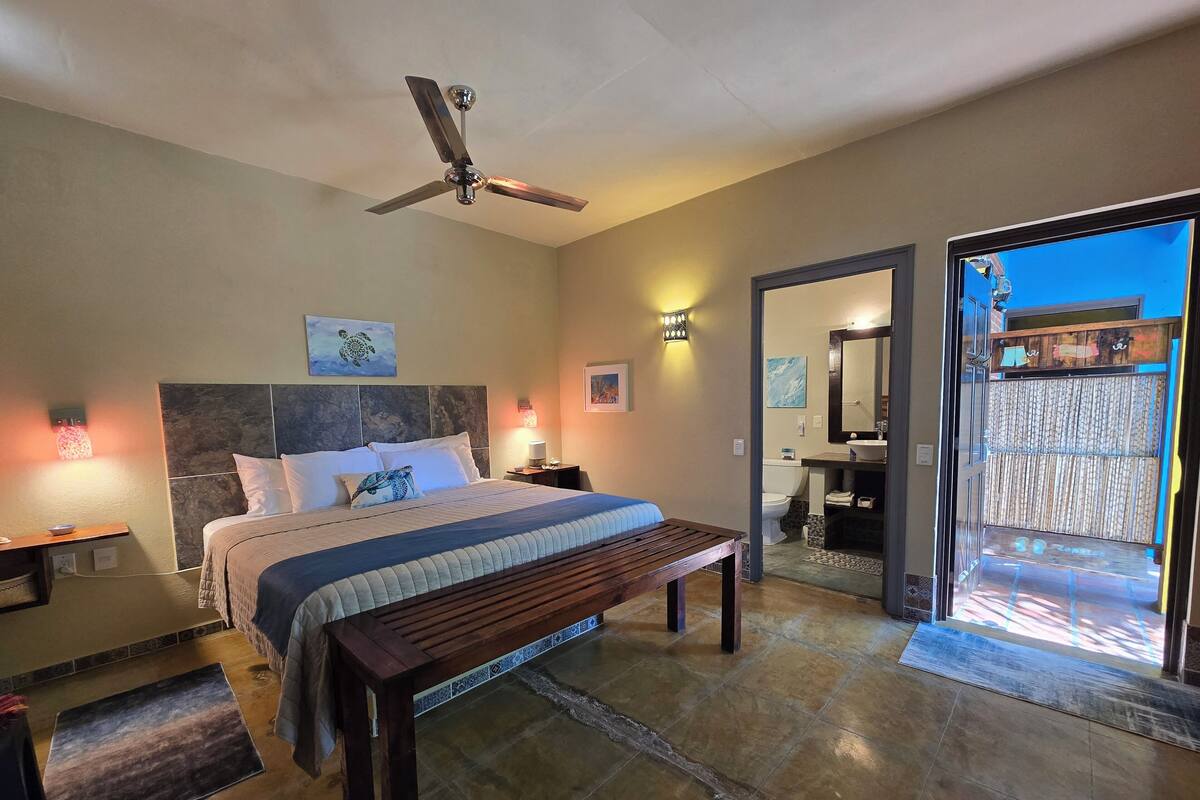 A hotel room with a bed, bedside table, bench, ceiling fan, and a view of the bathroom and outside.