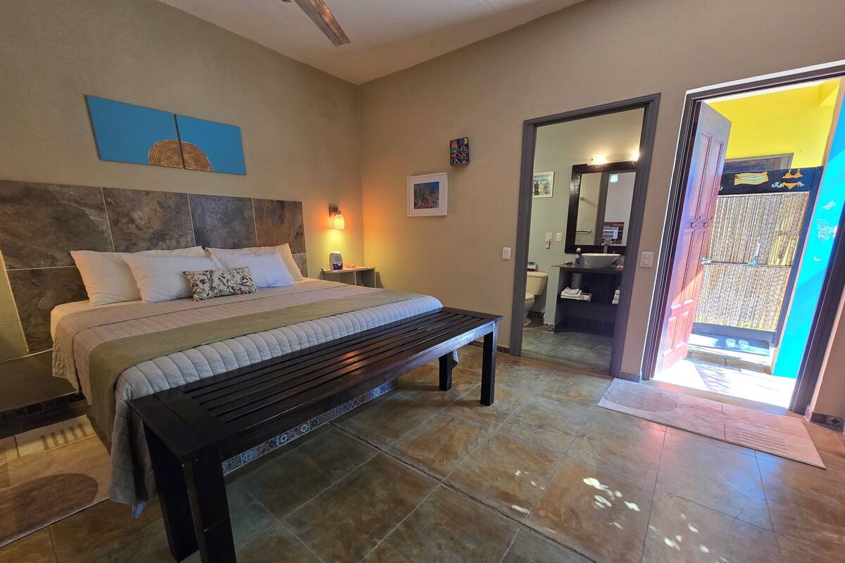 A bedroom with a bed, bedside tables, a bench, a wall-mounted artwork, and a bathroom visible through an open door.