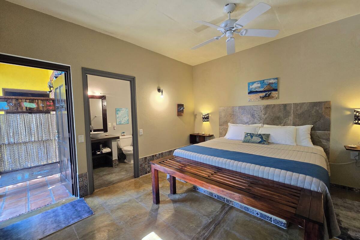 A bedroom with a bed, a ceiling fan, a nightstand, a wall-mounted lamp, and a bathroom visible through an open door.