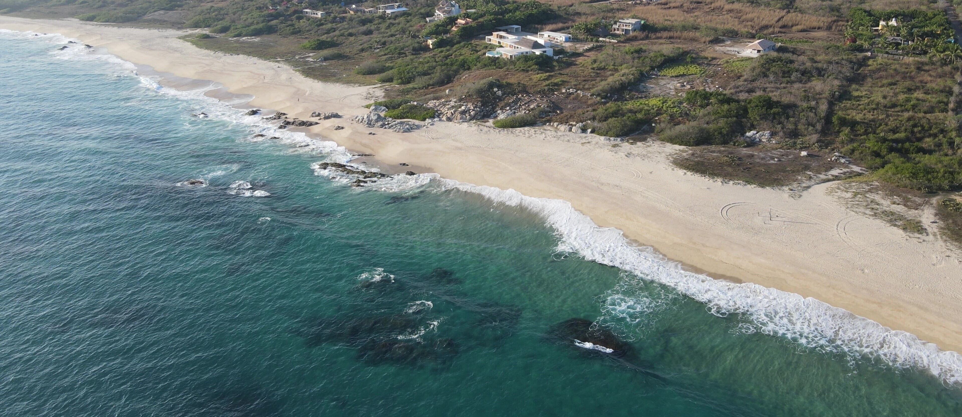 An aerial view of a beach with clear turquoise waters, sandy shore, and scattered buildings.