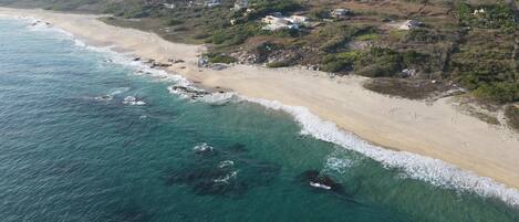 An aerial view of a beach with clear turquoise waters, sandy shore, and scattered buildings.