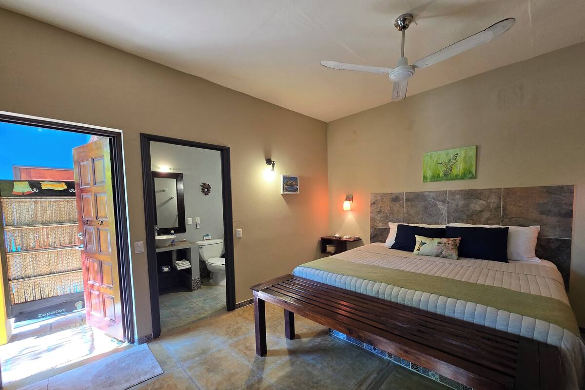 A bedroom with a bed, a ceiling fan, a nightstand, a wall-mounted light, and a view of the outside.