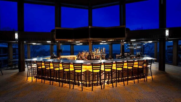 Una zona de bar con varios taburetes y una barra, ventanas amplias y vistas al exterior.