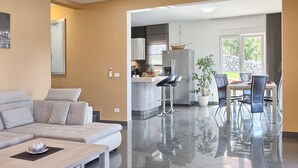 Ein modernes Wohnzimmer mit einer Couch und einem Couchtisch, eine Küche mit Kühlschrank und Barhocker sowie ein Essbereich mit Tisch und Stühlen.