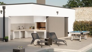 Un área de piscina moderna al aire libre con sillones de descanso, mesa de billar y una zona de cocina bajo un porche cubierto.