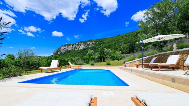 Una piscina con sillas de descanso y una sombrilla, rodeada de vegetación y una montaña al fondo.