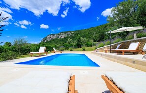 Une piscine entourée de chaises longues et d’un parasol, le tout bordé de verdure et avec une montagne en toile de fond.