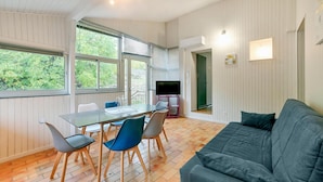 Ein modernes Wohnzimmer mit einem Glastisch, blauen und grauen Stühlen, einem dunkelgrauen Sofa und einem Fernseher auf einem rosa Sockel.