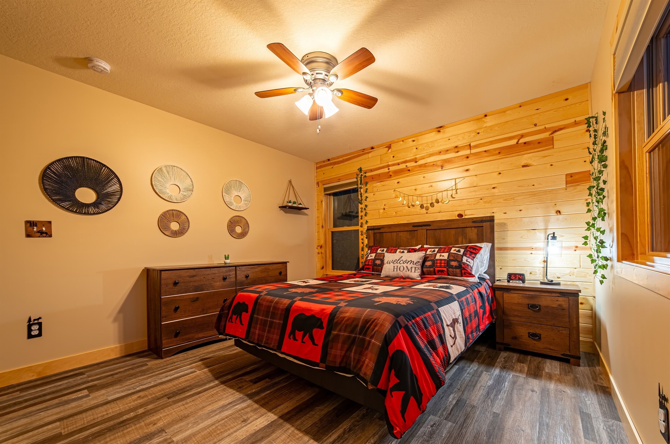 A bedroom with a bed, wooden headboard, ceiling fan, wall decorations, and a nightstand with a clock.