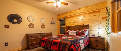 A bedroom with a bed, wooden headboard, ceiling fan, wall decorations, and a nightstand with a clock.