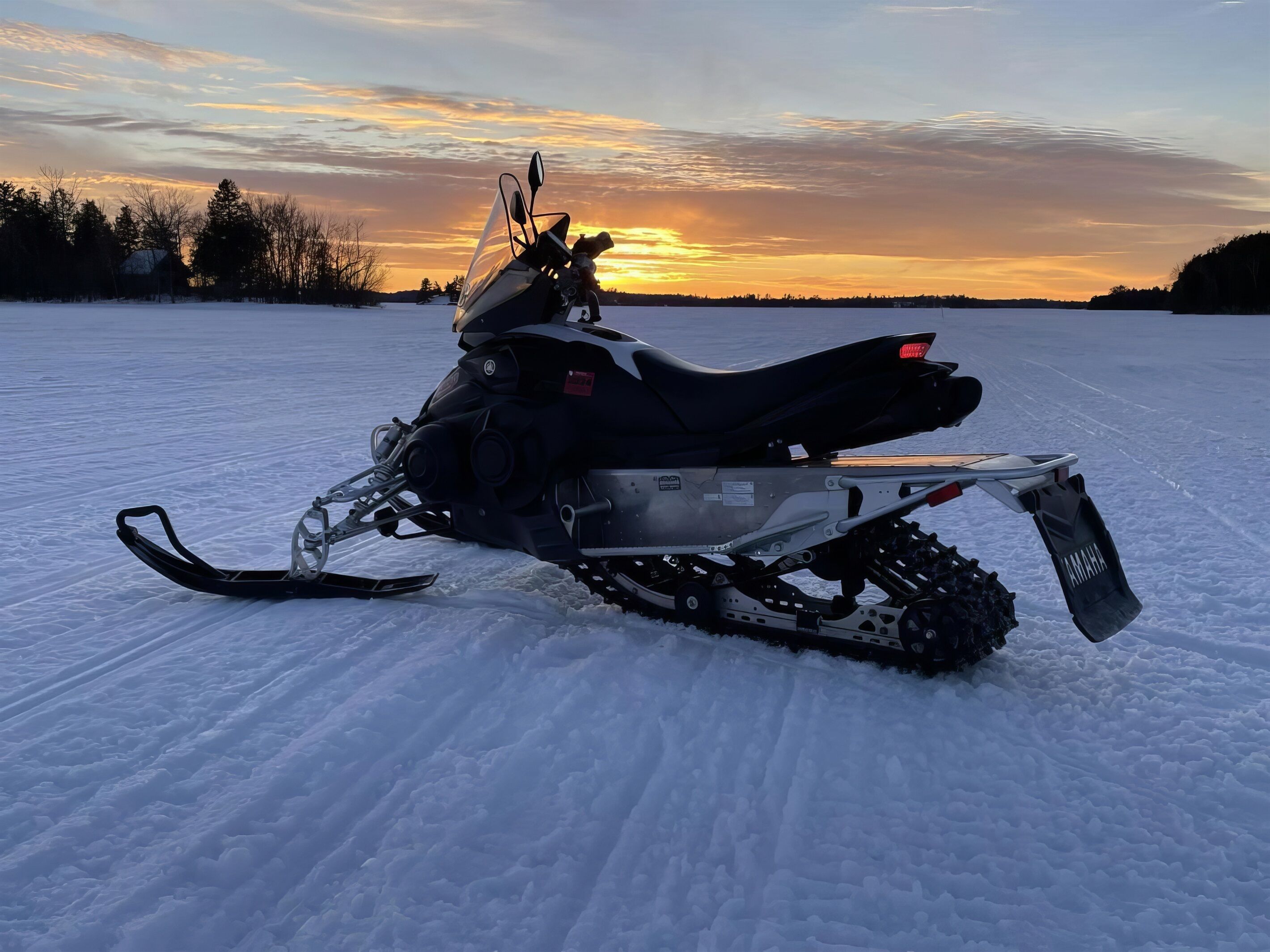 Una moto de nieve estacionada en un campo cubierto de nieve, con un atardecer de fondo.