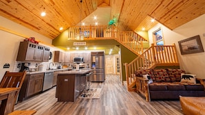 A spacious kitchen with wooden cabinets, a central island, and stainless steel appliances. A wooden staircase leads to a loft area with a green wall and a small Christmas tree. - Lake Views | Hot Tub | Games | Golf | Kayaks | St. Croix Casino (Turtle Lake)