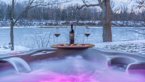 A hot tub with a plate of snacks and two glasses of wine, surrounded by trees and a frozen lake. - Lake Views | Hot Tub | Games | Golf | Kayaks | St. Croix Casino (Turtle Lake)