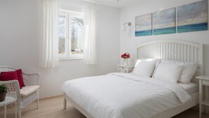 A bedroom with a large bed, a window with curtains, a white chair with a red pillow, a small table with a plant, and a wall with three paintings. - Cozy home in Labin - Rabac (Labin - Rabac)