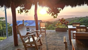 A wooden deck with a thatched roof, overlooking a scenic landscape with mountains and a sunset. - Ecohotel La Reserva (Piojó)