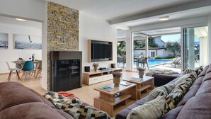A modern living room with a stone wall, a flat-screen TV, a dining area with a table and chairs, and a view of a swimming pool and outdoor seating. - Beautiful home in Pula (Pula)