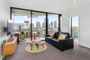 Un salon moderne avec un canapé en cuir noir, une petite table d’appoint en bois, un téléviseur fixé au mur et un balcon offrant une vue sur le paysage urbain.