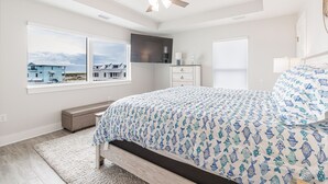 A bedroom with a large bed, a ceiling fan, a window with a view of houses, and a bedside table.