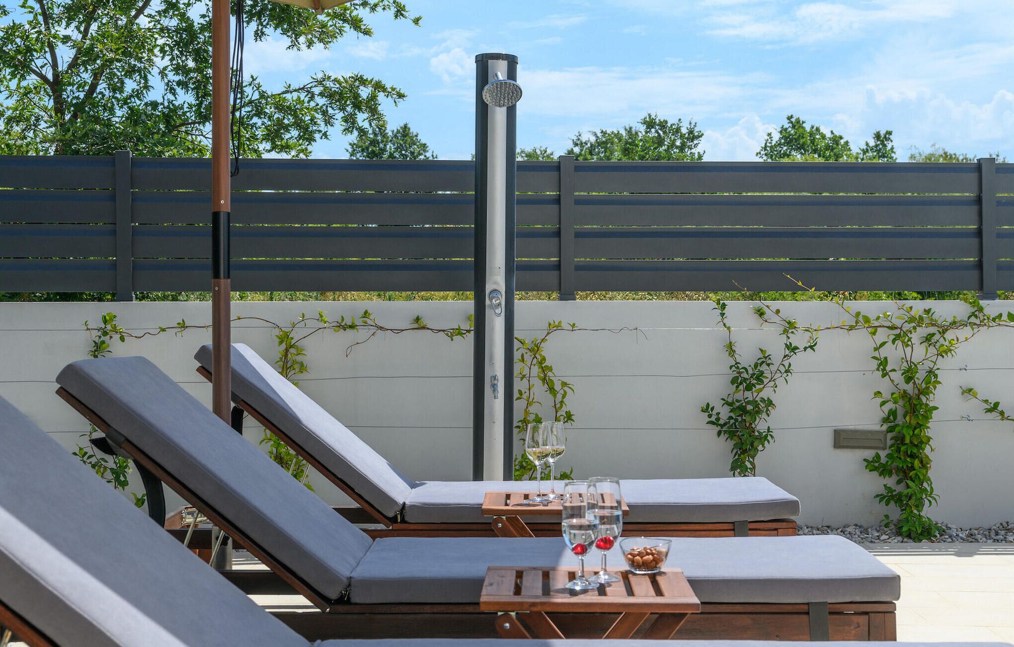 A sunlit outdoor lounge area with reclining chairs, a table with glasses and snacks, and a vertical water feature.