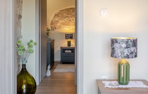 Ein Flur mit einer Steinsäule, einem grünen Strauß in einer Vase, einer Lampe mit floralem Lampenschirm und einem kleinen Tisch mit einer Spitzendecke.