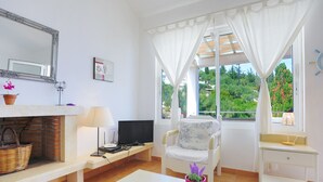 A bright living room with a fireplace, a television, a white sofa, and a window with curtains. - Dionisis Paxos Villa, 2-Story, Shared Pool, Sea Views, Modern Amenities (Yioliskari)
